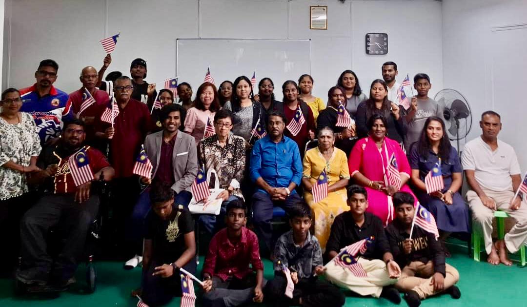 Adults and children at a community video course in Johor pose together holding Jalur Gemilang flags ahead of Merdeka.